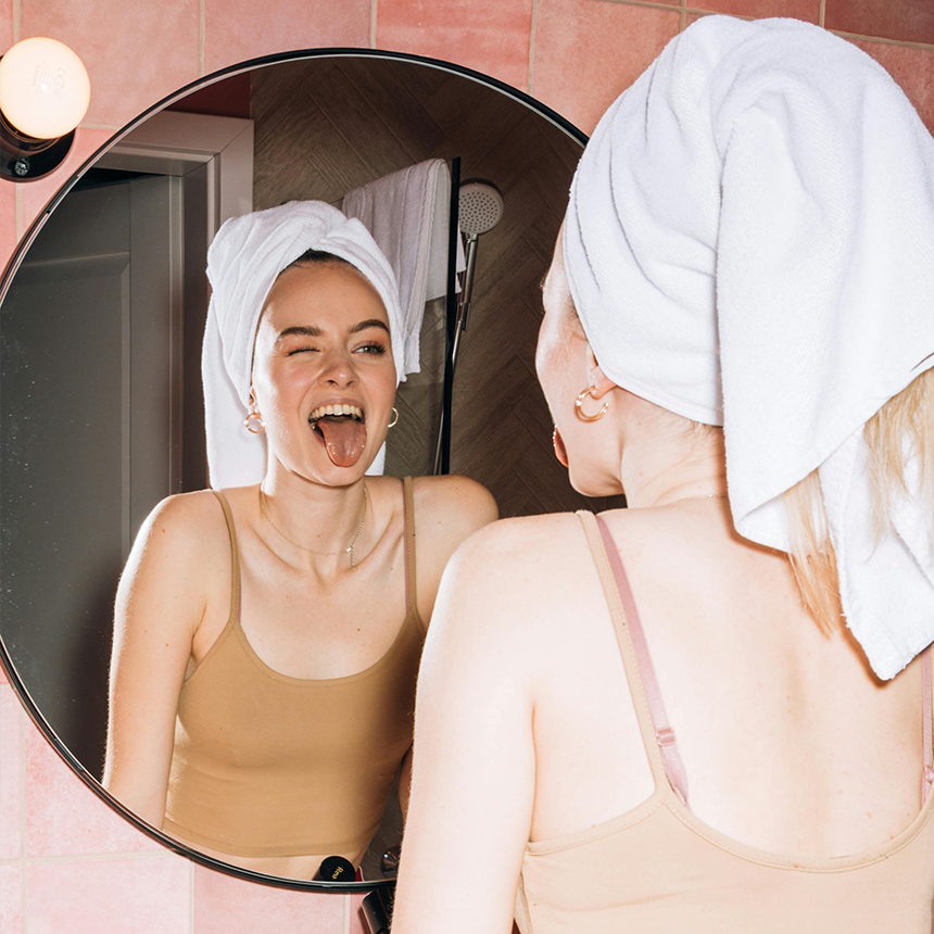 Junge Frau mit einem Handtuch um den Kopf gewickelt, schaut in den Spiegel und streckt sich selber die Zunge raus