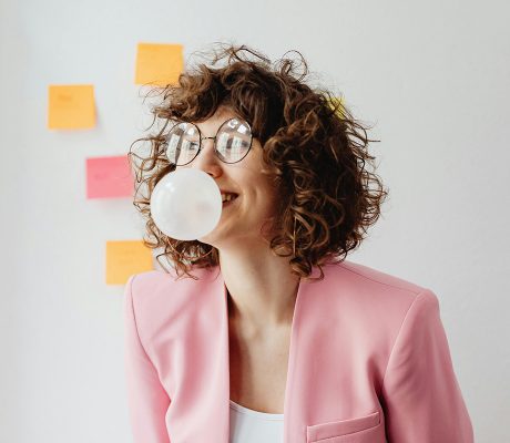 Frau in rosa Blazer mit lockigem Haar und einer Brille lacht, während sie eine Blase mit einem Kaugummi wirft