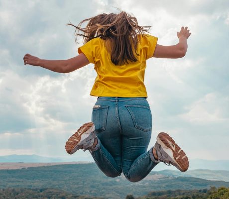 Frau springt in die Luft. Man sieht sie von hinten und im Hintergrund ist eine hügelige Landschaft zu sehen